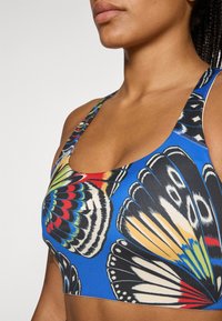 Αθλητικό σουτιέν με μοτίβο μπλε πεταλούδες, με στρογγυλό λαιμό, πολύχρωμες λεπτομέρειες και κεντημένο λογότυπο στη μέση. Μαλακό και ελαστικό ύφασμα.