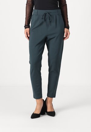 Woman wearing teal tapered pants with a drawstring waist and black high-heeled shoes standing on a white background.