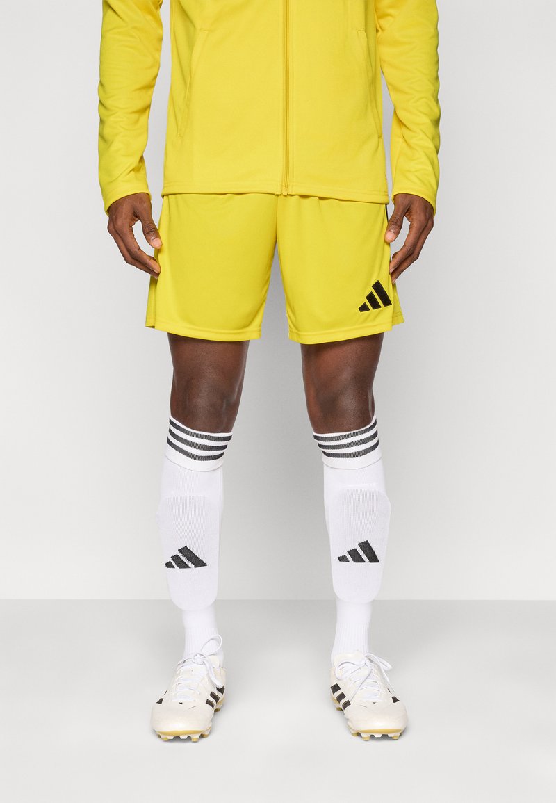 Σετ αθλητικής ένδυσης σε κίτρινο χρώμα, που περιλαμβάνει ζακέτα με φερμουάρ και σορτς, με μαύρα λογότυπα Adidas. Λευκές κάλτσες με μαύρες ρίγες και ποδοσφαιρικά παπούτσια.