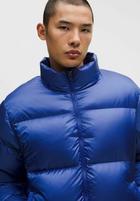 Sinine puff-jakk kõrge kraega, millel on läikiv tekstuur, tõmblukk ja nähtavad horisontaalsed õmblused isolatsiooni jaoks.