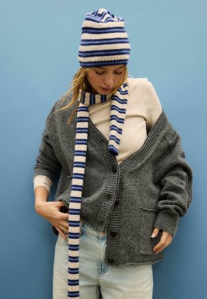 Sivi pleteni kardigan z gumbi in žepi, kombiniran s črtasto modro-belo kapo in šal. Model nosi svetel top in kavbojke.