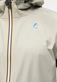 Μπεζ μπουφάν K-Way με ρίγα φερμουάρ σε λευκό, ναυτικό μπλε και κίτρινο, διαθέτει κουκούλα και το λογότυπο K-Way στο στήθος.
