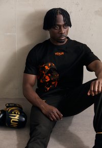 Sort bomuld t-shirt med orange camouflage-mønster og "VENUM" logo. Sorte atletiske bukser ved siden af sorte og guld boksehandsker.