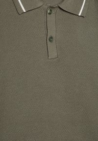 Alyvuogių spalvos polo marškiniai su tekstūruotu mezgimo raštu, dviem tamsiomis sagomis ir apykakle, papuošta plonomis baltomis juostelėmis.