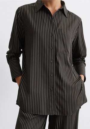 Zwart overhemd met knoopsluiting en bijpassende broek met dunne witte verticale strepen gedragen door een persoon met handen in de zakken.