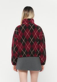Rød og svart argyle-mønstrete fleece-genser med høy krage, cropped design og myk tekstur. Kombinert med en grå miniskjørt.