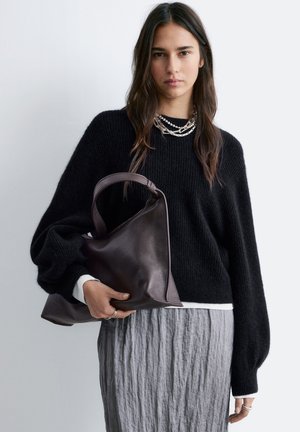 Ung kvinde iført en sort sweater og grå nederdel holder en stor mørkebrun lædertaske med lagdelte sølvsmykker.