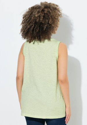 Persoon met krullend haar die een mouwloos lichtgroen topje en donkerblauwe broek draagt, gericht naar een witte muur, van achteren afgebeeld.