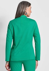 Grüner, taillierter Blazer aus glattem Stoff mit einem maßgeschneiderten Schnitt, langen Ärmeln und subtilen Nahtdetails. Kombiniert mit passenden Hosen.