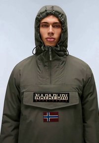 Mlad oseba nosi olivno zeleno jakno s kapuco, na kateri je logo Napapijri in našitek norveške zastave na sprednjem žepu.