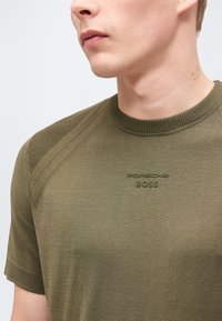 Νεαρός άνδρας που φοράει ελαφρώς πράσινο πλεκτό πουκάμισο με κοντά μανίκια και ανάγλυφο λογότυπο "Porsche BOSS" στο στήθος.