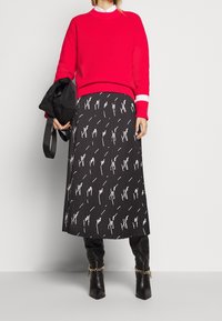 Pull rouge côtelé avec des rayures blanches aux manches, associé à une jupe noire avec un motif abstrait blanc, et des bottes noires montantes.