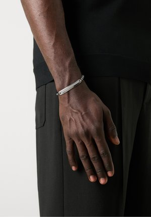 Bracelet en argent avec un design en barre texturée et des accents en matériel ronds, porté sur une main contre des vêtements noirs.