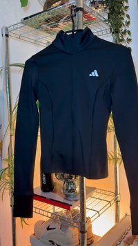 Crna sportska jakna dugih rukava s visokim ovratnikom, s bijelim logotipom, prilagođenim dizajnom i detaljima na šavovima.