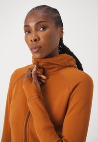 Orange zip-up hoodie med hög krage, i mjukt tyg och med tumhålsknappar, har en framträdande logotyp nära huvan.