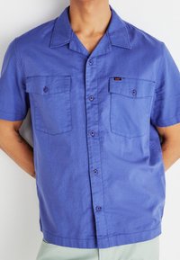 Kortärmad blå skjorta i lättviktstyg, med krage, två bröstfickor och kontrasterande knappräder.