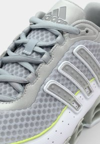 Svetlo sivi atletski copat z mrežasto zgornjo plastjo, tremi belimi črtami in izrazitim belim podplatom, okrašenim z rumeno. Ima večplastno zasnovo.