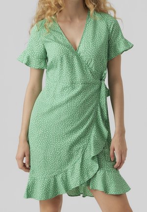 Femme portant une robe portefeuille verte à pois blancs, manches flottantes, ourlet à volants et fermeture à nouer sur le côté, sur fond gris.