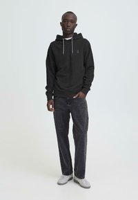 Zwarte hoodie met trekkoorden, met een logo op de borst, gecombineerd met donkergrijze spijkerbroek en grijze sneakers. Effen achtergrond.