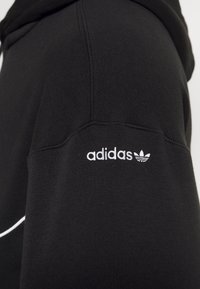 Le sweatshirt noir présente une capuche, des manches longues et un logo "adidas" brodé en blanc avec le trèfle sur l'épaule gauche. Texture douce.