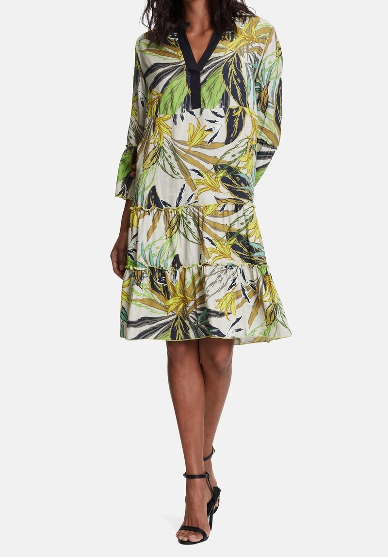 Robe à manches courtes avec un motif de feuilles tropicales dans des teintes jaunes, vertes et noires. Elle présente un décolleté en V et un design à volants superposés.