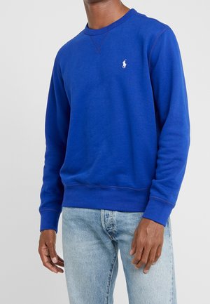 Mann trägt ein leuchtend blaues Langarm-Sweatshirt mit kleinem weißem Polo Ralph Lauren-Logo, kombiniert mit hellblauen Jeans.