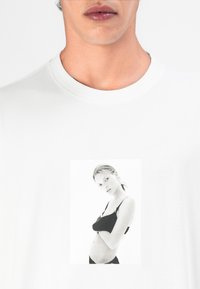 Witte T-shirt met een zwart-wit fotoprint van een model in een zwarte bh. Katoenen stof, ronde hals, korte mouwen.