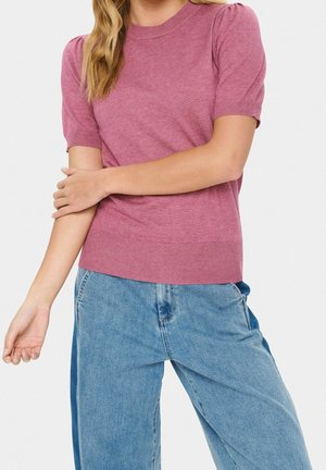 Kvinde iført en lyserød kortærmet top og løstsiddende lyseblå jeans med armene krydset om livet på en ensfarvet baggrund.