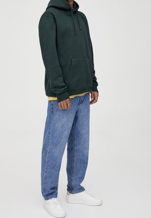 Felpa verde scuro con cappuccio e tasca frontale, abbinata a jeans larghi azzurri chiari e scarpe da ginnastica bianche. Design semplice, senza motivi visibili.