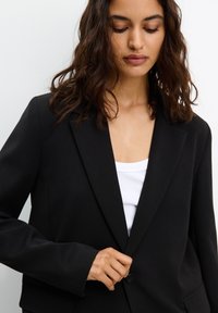 Čierny blazer s klopovým golierom, s jedným gombíkom a prednými vreckami. Nosený cez biely top bez rukávov, s hladkým povrchom.