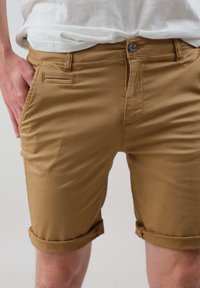 Shorts en khaki en coton léger, dotés d'un bouton et d'un zip, de quatre poches et de poignets roulés. Texture lisse sans motifs.