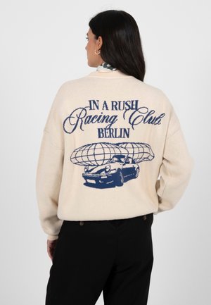 Kremfarget genser med oversized passform, med marineblått motiv av en bil og teksten "IN A RUSH Racing Club BERLIN" på baksiden. Myk tekstur.