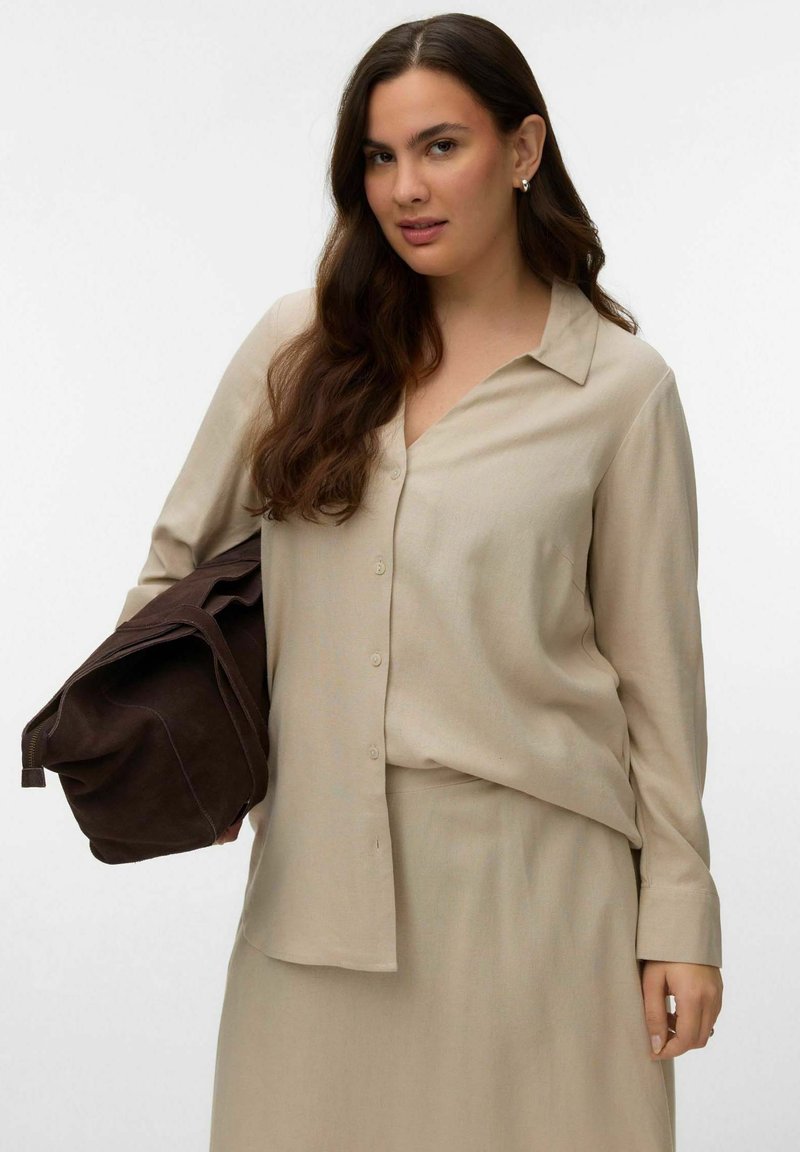 Femme aux longs cheveux bruns portant une chemise beige boutonnée et une jupe assortie, tenant un grand sac en cuir marron foncé sur son bras.