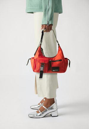 Borsa in nylon rossa con molte tasche, una fibbia argentata e accenti neri, tenuta accanto a pantaloni beige e tacchi alti argentati lucidi.