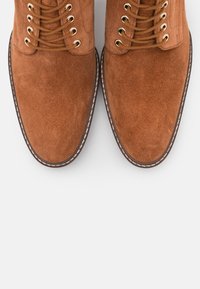 Stivali in suede marrone con occhielli dorati, lacci rotondi e una finitura testurizzata. La cucitura di contrasto delineano la suola e la zona della punta.