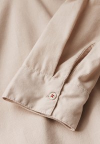 Puño de camisa color beige claro con textura de rayas verticales, que presenta un botón con un diseño floral en rojo y blanco.