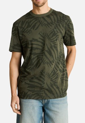 Homme portant un t-shirt vert à manches courtes avec un motif feuillu foncé et un jean bleu clair, debout devant un fond uni.
