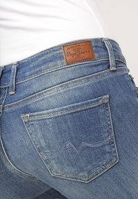 Blå denimjeans med en läderpatch i midjan. Har en böjd bakfickdesign med sömdetaljer. Tyget har en något sliten textur.