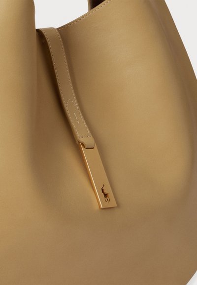 Bež kožna torba za nošenje s glatkom teksturom, s ravnim remenom i pravokutnim zlatnim ukrasom s utisnutim logotipom životinje.