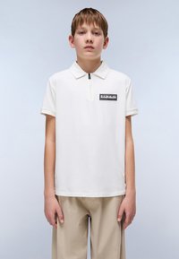 Hvit polo-skjorte med halv glidelås og en svart rektangulær logo-patch på venstre bryst, kombinert med beige bukser. Glatt bomullsstruktur.
