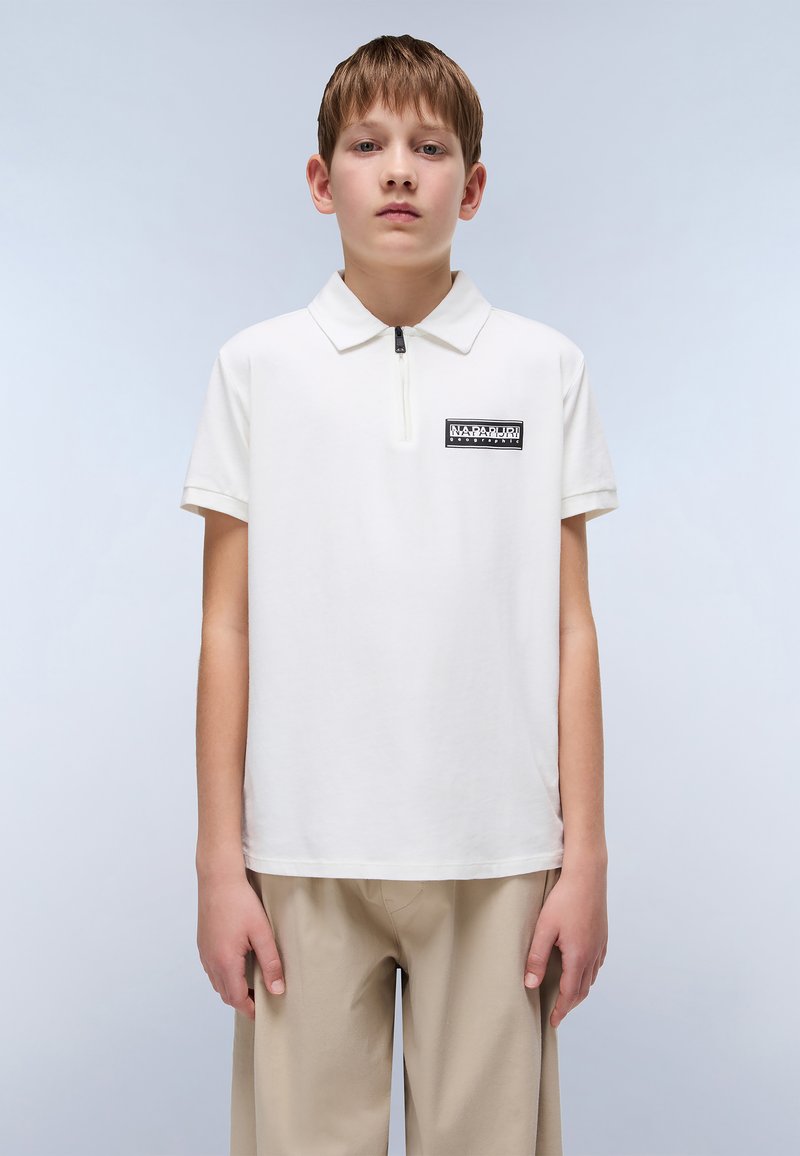 Hvit polo-skjorte med halv glidelås og en svart rektangulær logo-patch på venstre bryst, kombinert med beige bukser. Glatt bomullsstruktur.