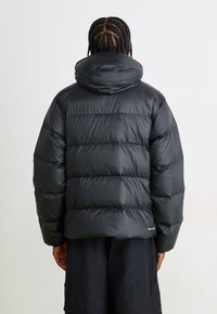 Sort pufferjakke med hætte, opdelt i quiltede sektioner. Har en glat, blank finish og elastiske manchetter. Minimal branding synlig.