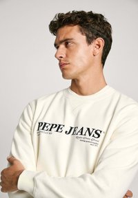 Krémová mikina s černým logem "PEPE JEANS", kulatým výstřihem a lemováním na rukávech. Model stojí s překříženýma rukama na neutrálním pozadí.