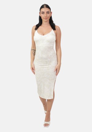 Frau mit dunklen Haaren, die ein eng anliegendes weißes Midikleid mit Blumenmuster und weiße High Heels trägt, geht vor einem schlichten hellen Hintergrund nach vorne.