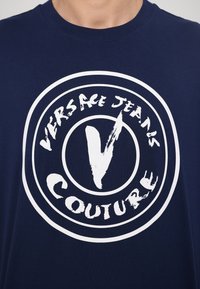 Marineblå bomulds T-shirt med en cirkulær hvid logo med teksten "VERSACE JEANS COUTURE" og et fremtrædende stiliseret "V" i midten.