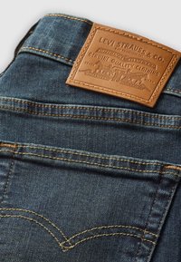 Poche arrière d'un jean en denim bleu foncé avec surpiqûres jaunes et un patch en cuir beige Levi Strauss & Co. avec logo et texte embossés.