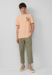 Lichtoranje katoenen T-shirt met korte mouwen en een bedrukt ontwerp; gecombineerd met olijfgroen, relaxed-fit broeken en beige sneakers.