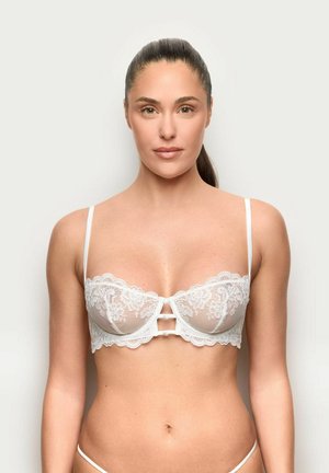 Kobieta z włosami zaczesanymi do tyłu, ubrana w biały koronkowy biustonosz typu balconette, na jednolitym jasnym tle, zwrócona przodem z neutralnym wyrazem twarzy.