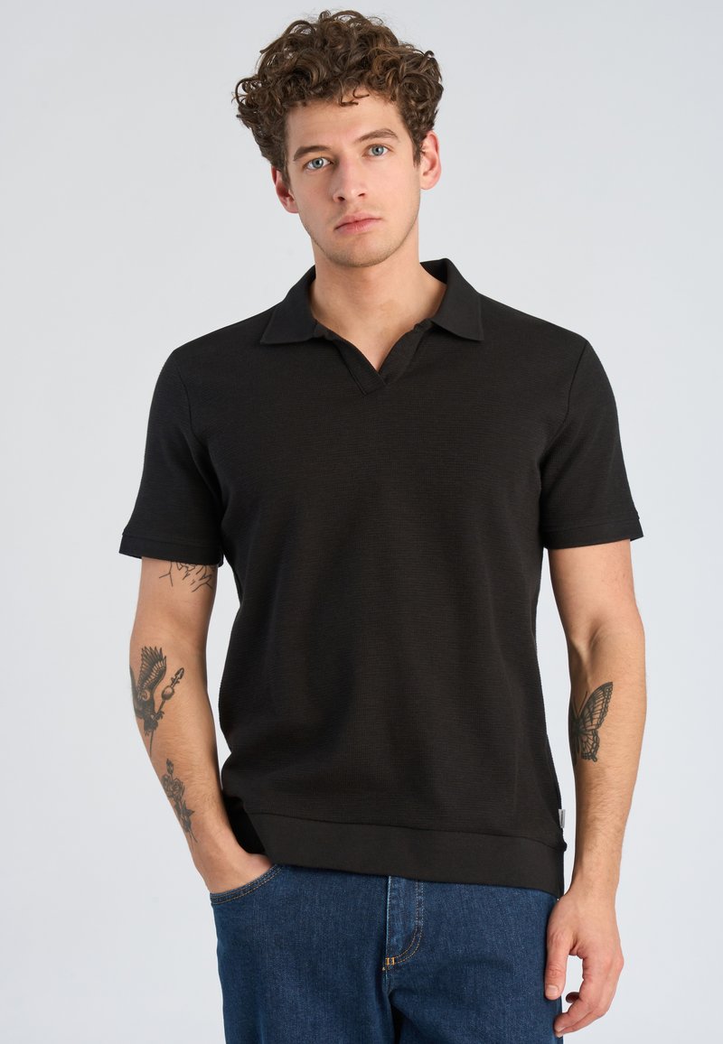 Junger Mann mit lockigem Haar, trägt ein schwarzes kurzärmeliges Poloshirt und blaue Jeans, zeigt Tätowierungen auf beiden Unterarmen, steht vor einem grauen Hintergrund.