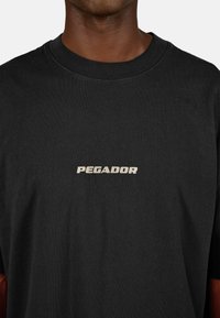 Czarny bawełniany t-shirt z okrągłym dekoltem. Na klatce piersiowej znajduje się napis "PEGADOR" w kontrastującym jasnym kolorze. Tkanina wydaje się gładka.